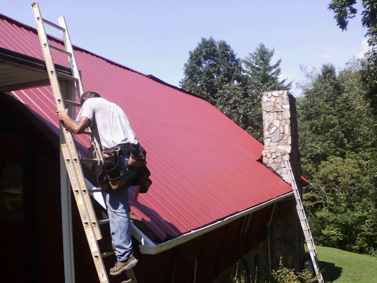 Roofer on a ladder beside a red metal roof and stone chimney — Soffit & Fascia Repair by Roofing Brothers in Millersville